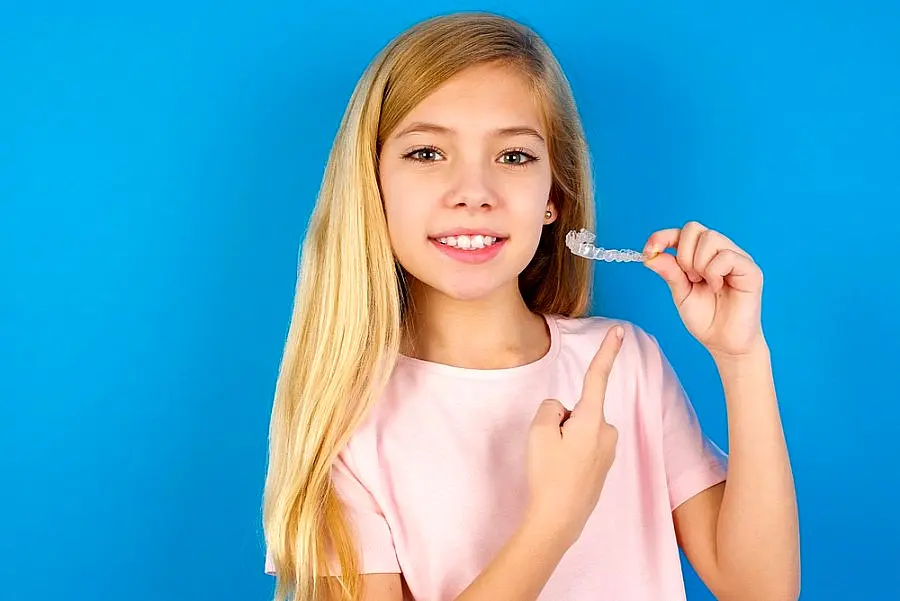 Young girl holding and pointing at a clear dental aligner against a blue background, wearing a pink shirt in Sammamish or Kent, WA.