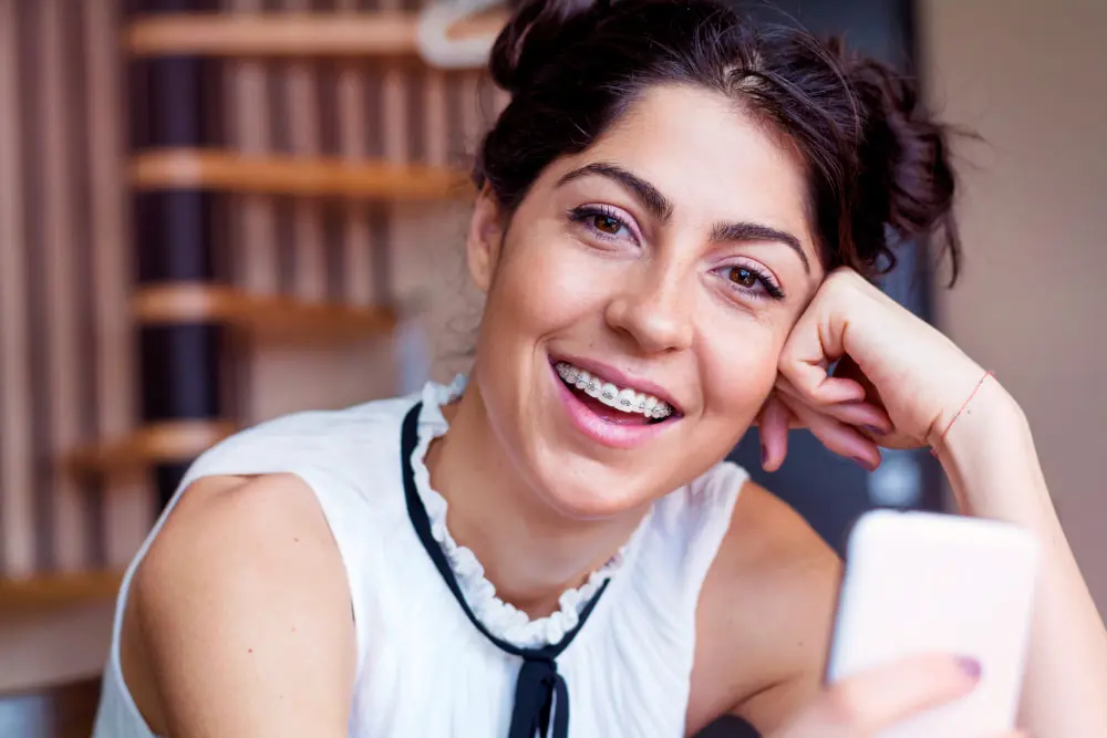A person with braces, wearing a white shirt, is smiling and holding a smartphone indoors. Illustrating adult braces at Freelove Orthodontics in Sammamish and Kent, WA