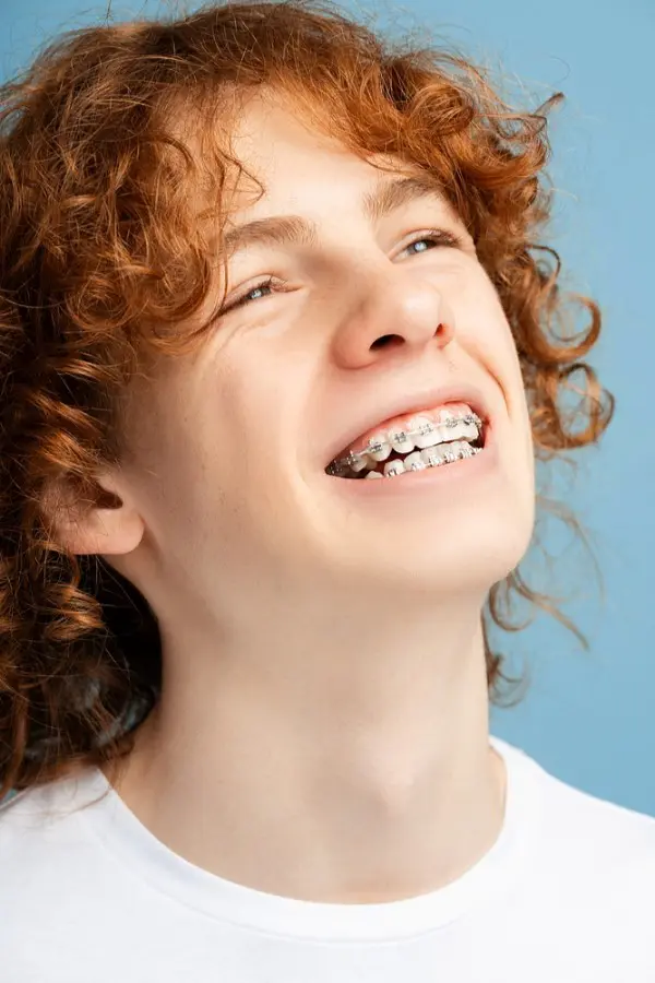 Smiling person with curly red hair and metal braces from orthodontist in Sammamish, WA, wearing a white shirt against a blue background.
