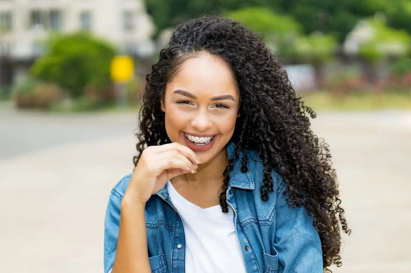 A person with long, curly hair and metal braces smiles outdoors near orthodontist in Sammamish Kent, WA, wearing a denim jacket over a white shirt.