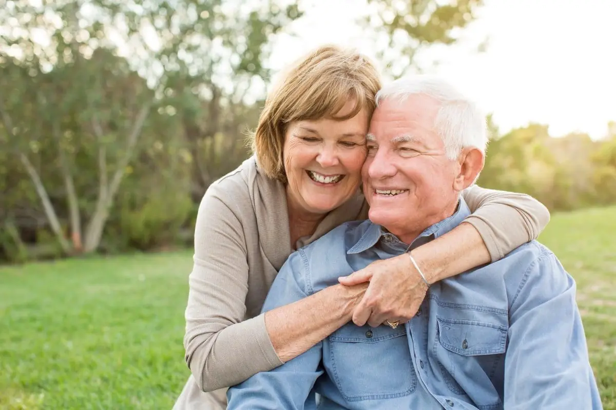An elderly couple sits on the grass, the woman hugging the man from behind. Despite their age, they smile brightly, surrounded by greenery—a testament to happiness being life's best braces or orthodontic insurance in Sammamish or Kent, WA.