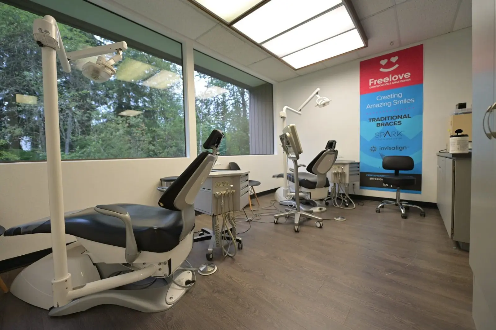 Orthodontist in Sammamish or Kent, WA office with two treatment chairs, modern equipment, and a large window overlooking the vibrant community. A banner showcases various dental services. The room features elegant wood flooring and ceiling lights for a welcoming ambiance.