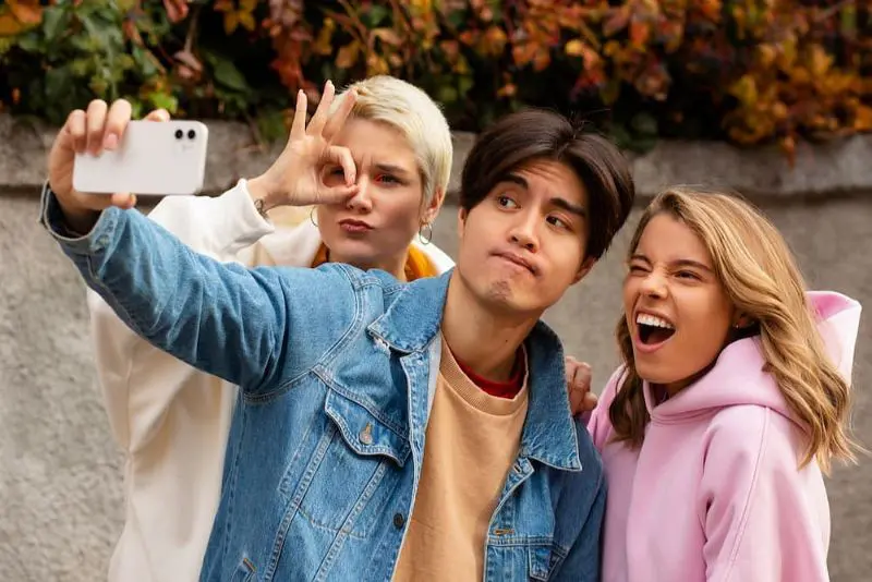 Three people pose for a playful selfie outdoors. One holds the phone, another gestures an 'OK' sign, and the third smiles widely. They stand in front of a wall with autumn leaves. Illustrating Types of Braces in Sammamish and Kent WA