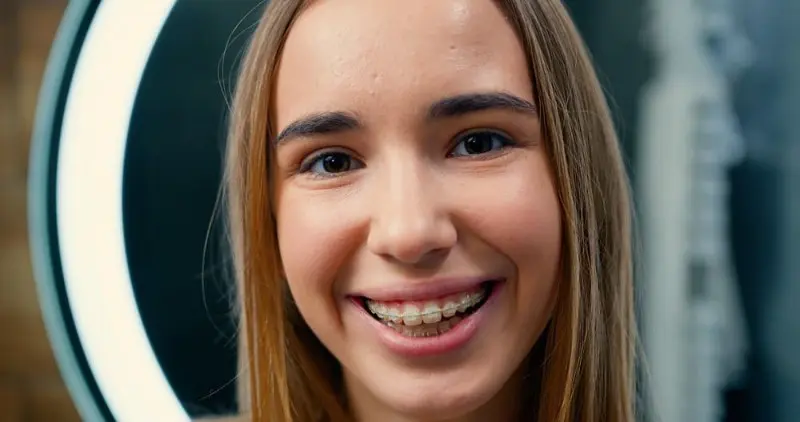 A young person with long hair, ceramic clear braces and a radiant smile, stands confidently in front of a ring light, capturing the result of orthodontist in Sammamish, WA.