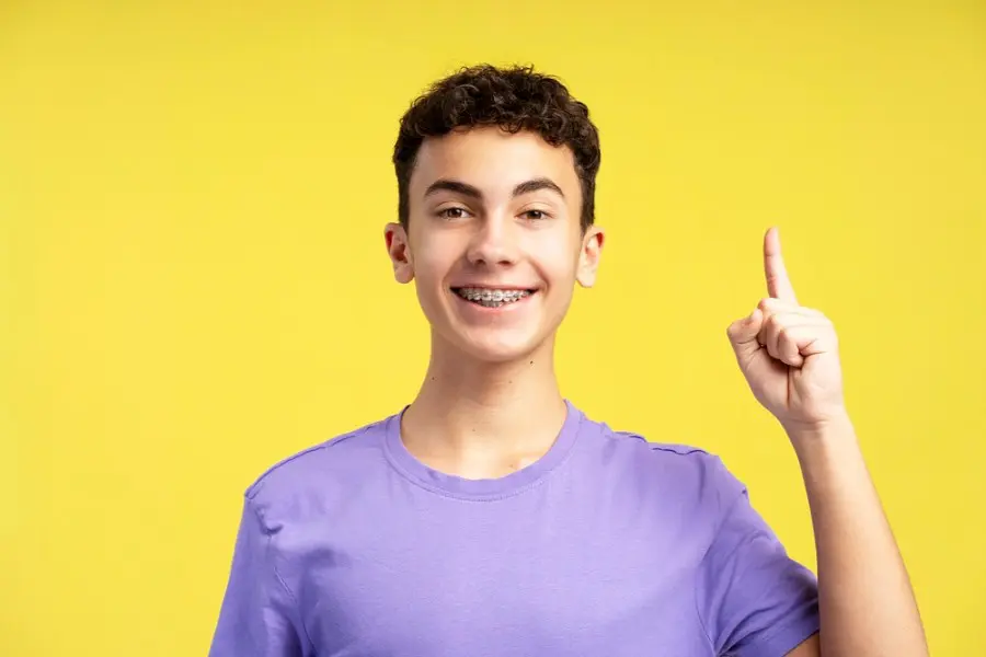 Teenager in a purple shirt with braces smiles, raising one finger against a bright yellow background in Sammamish or Kent, WA.