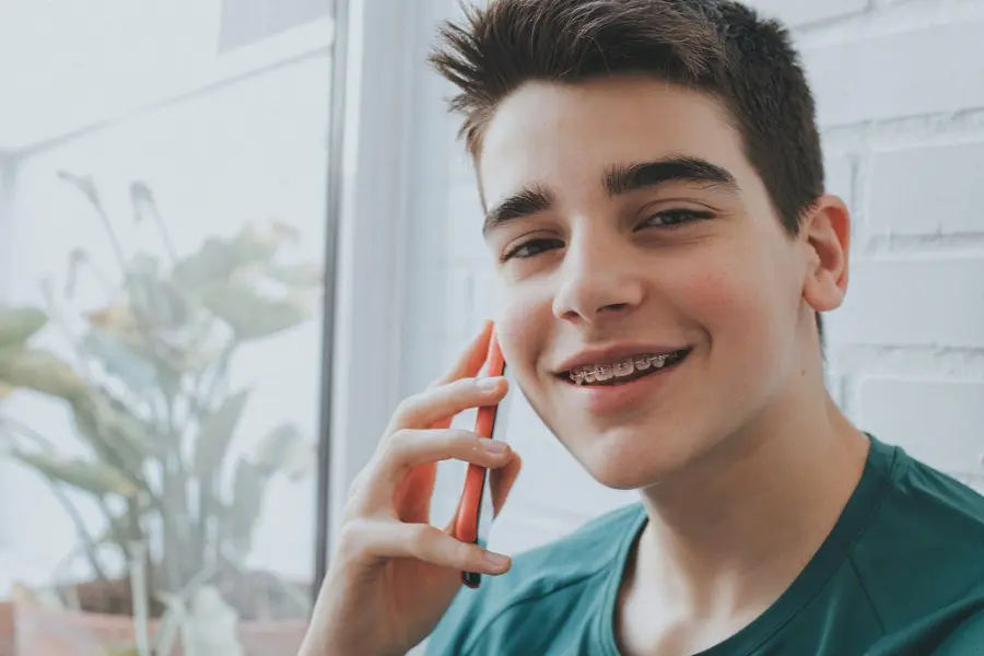 Teenage boy with braces smiling and talking on a red phone indoors, next to a window with plants in Sammamish or Kent, WA.