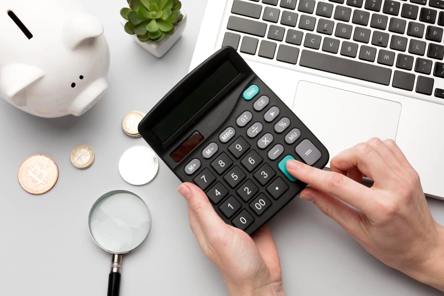 Hands using a calculator on a table with a laptop, piggy bank, coins, and magnifying glass highlight the importance of financial planning. A small plant represents growth as orthodontic insurance plans ensure your smile investment flourishes alongside your savings in Sammamish or Kent, WA